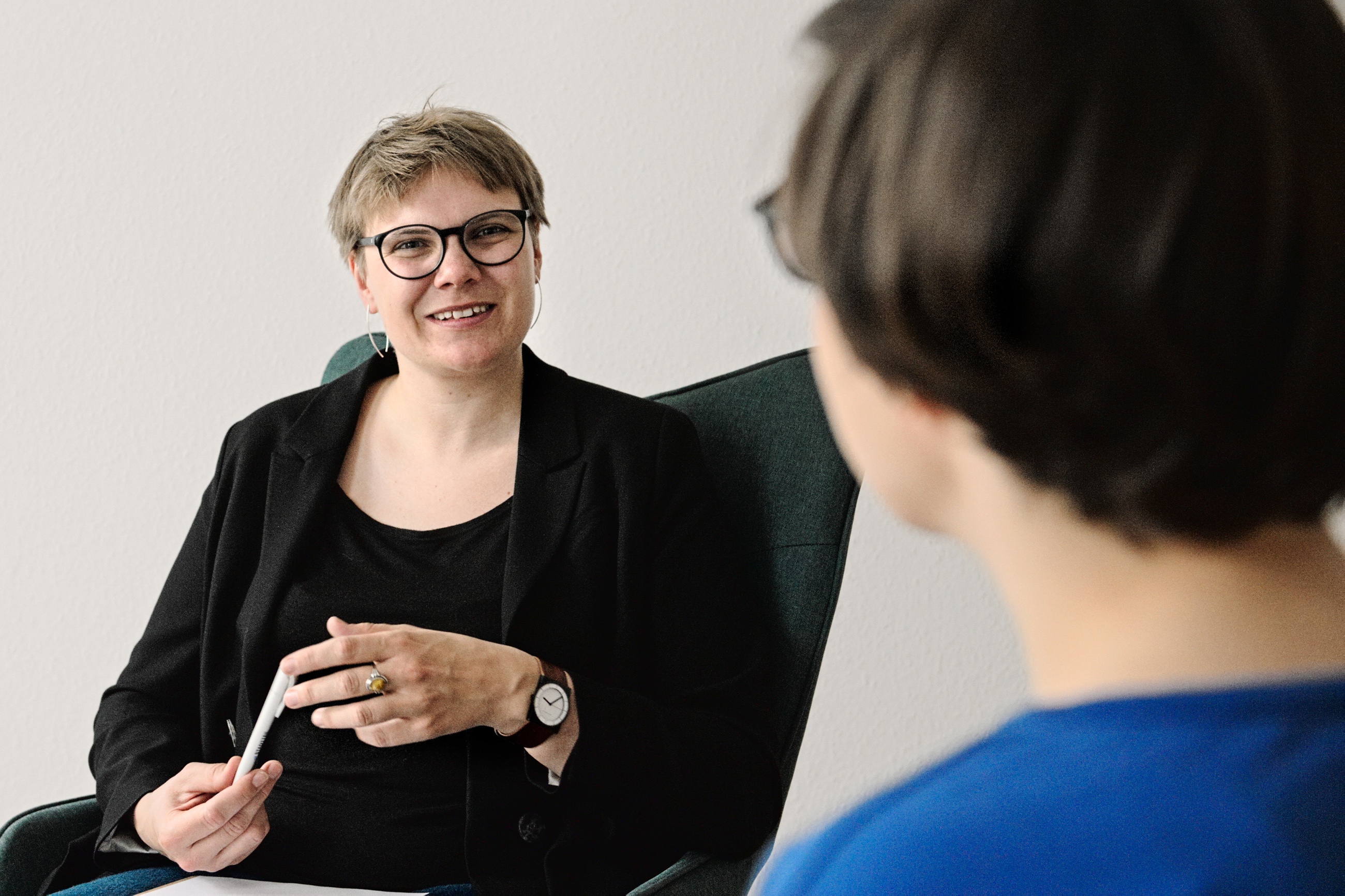 Luise Veit ist im Coaching mit einer Klientin. Es ist der Oberkörper von zu Luise Veit sehen. Sie sitzt mit einem schwarzen Blazer bekleidet in einem Sessel. Luise Veit hat kurze Haare, eine Brille mit schwarzem Rand und trägt silberne Ohrringe. Sie blick