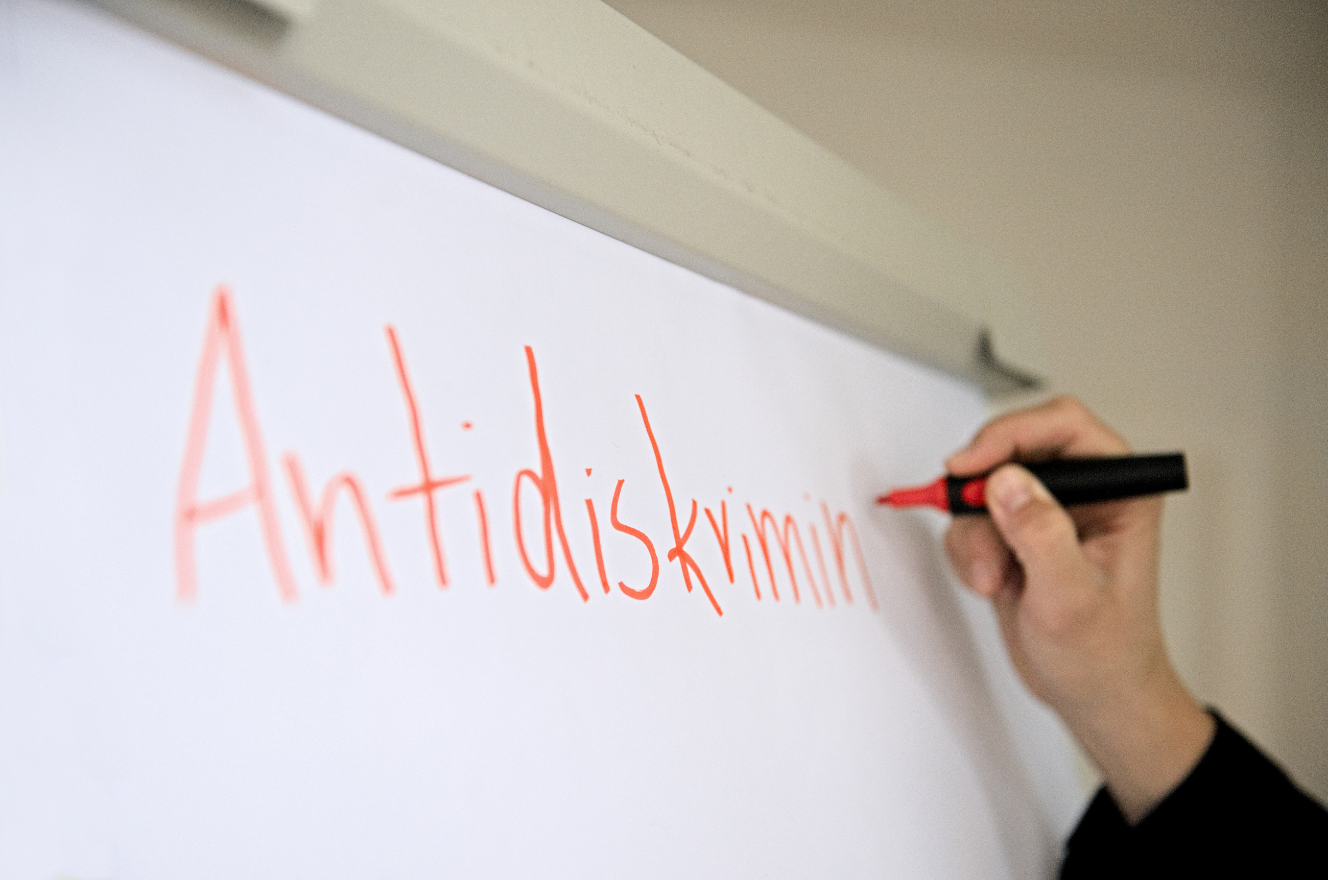 Auf einem weißen Flipchart steht etwas verschwommen „Antidiskrimin“ in oranger Schrift. Nach dem letzten Buchstaben ist ein Moderationsstift der Marke Neuland in orange zu sehen. Er wird von der Hand von Luise Veit geführt.