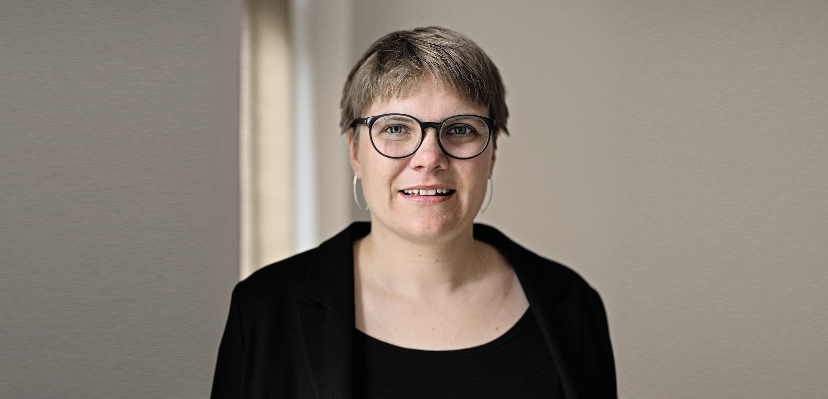 Portrait von Luise Veit. Sie lacht freundlich nach vorn. Luise Veit trägt kurze Haare, eine Brille mit dunklem Rand und zwei silberne Ohrringe. Sie trägt ein schwarzes Oberteil und einen schwarzen Blazer. Im Hintergrund ist eine helle Wand.