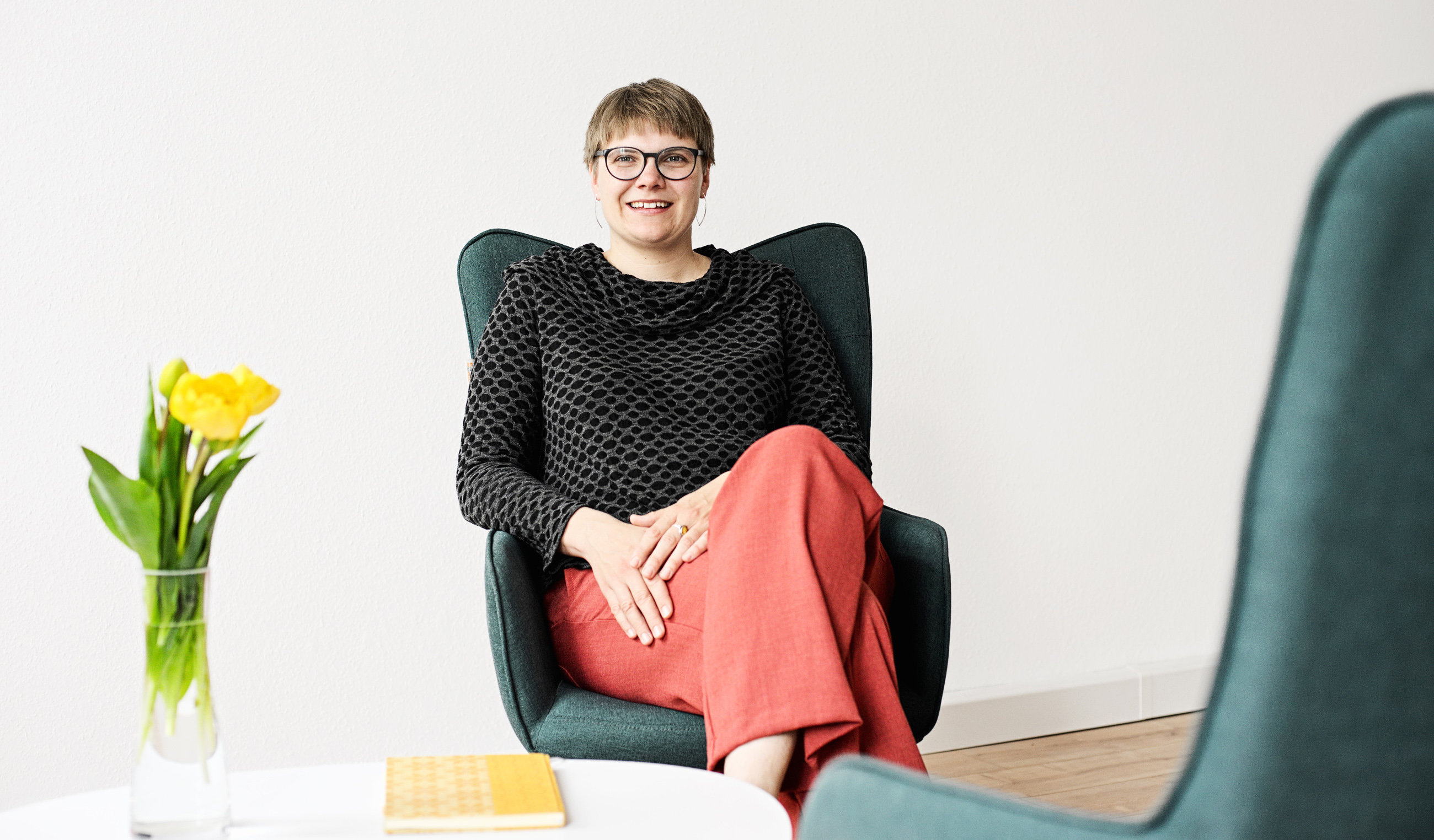 Luise Veit sitzt in einem dunkelgrünen Ohrensessel. Sie trägt kurze Haare, eine Brille mit dunklen Rand, silberne Ohrringe, einen schwarz-grauen Rollkragenpullover und eine rote Hose. Ihre Beine sind überschlagen. Sie blickt freundlich nach vorn. Vor ihr 