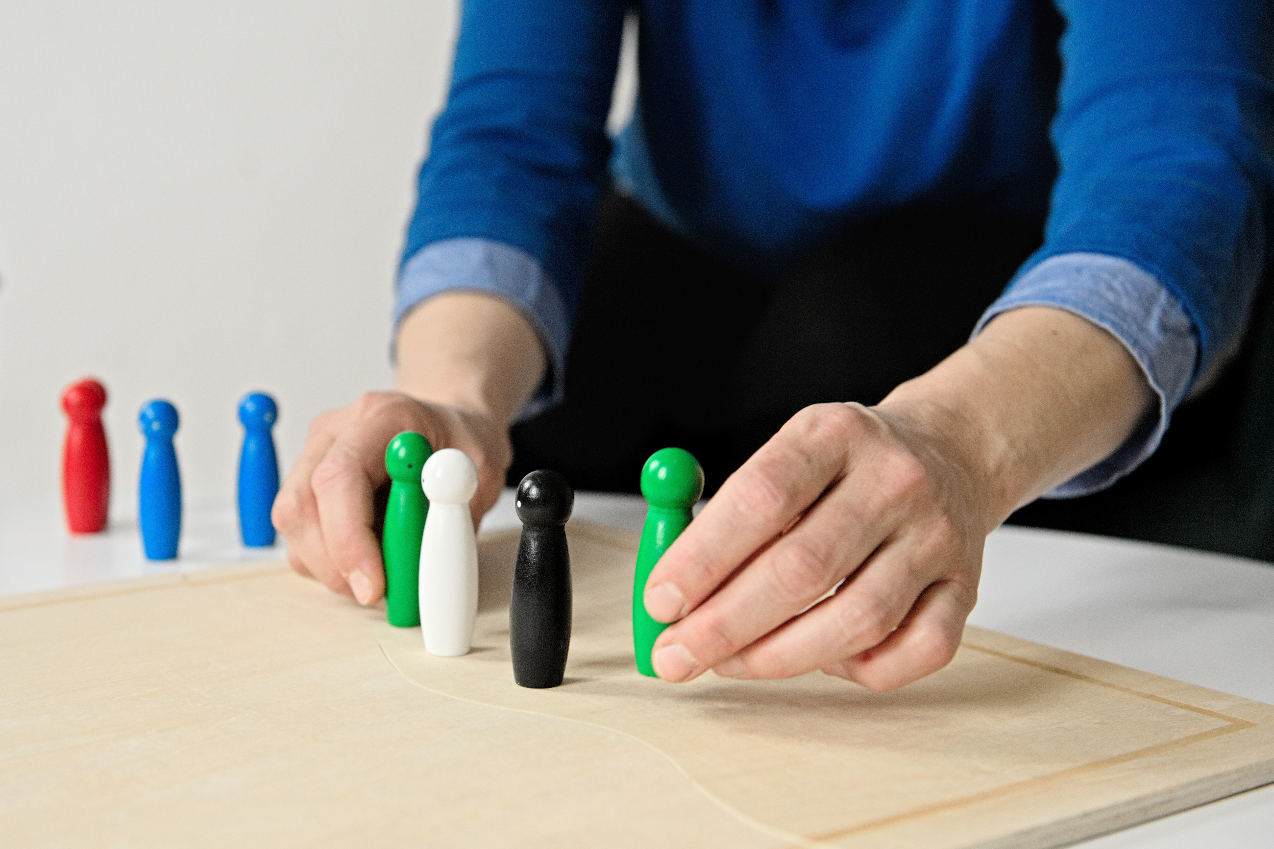 Auf einem weißen Tisch liegt ein hellbraunes Systembrett aus Holz. Auf dem Brett stehen zwei grüne, eine weiße und eine schwarze Figur. Die Hände von Luise Veit umfassen die grüne Figur, die jeweils links und rechts der schwarzen und weißen Figuren stehen