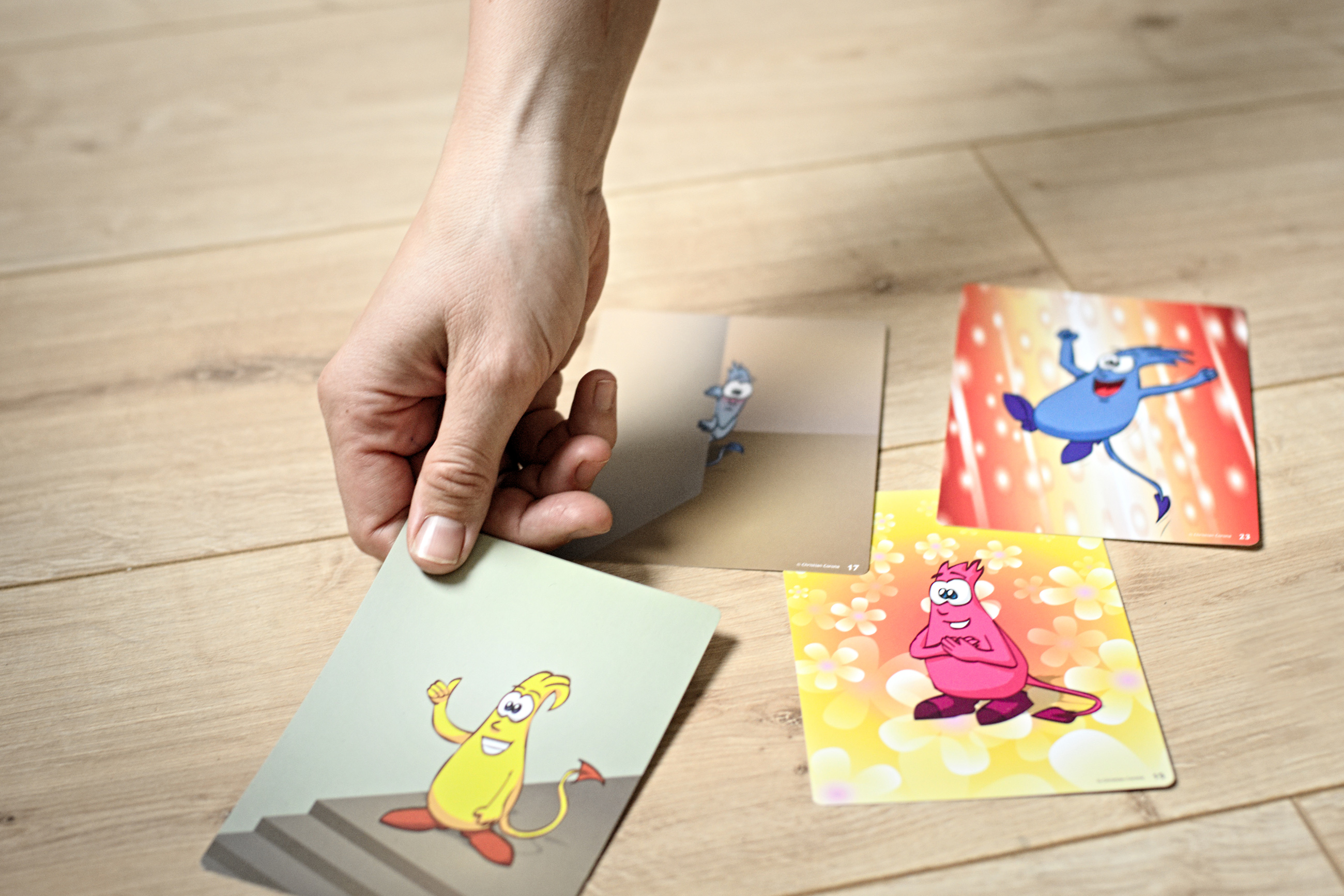 Eine Hand legt auf einen Holzboden sogenannte Monsterkarten. Das sind Moderationskarten mit Monstern, die verschiedene Emotionen zum Ausdruck bringen. Die eine Karte zeigt ein gelbes Monster mit Daumen hoch am oberen Ende einer Treppe. Auf einer anderen K